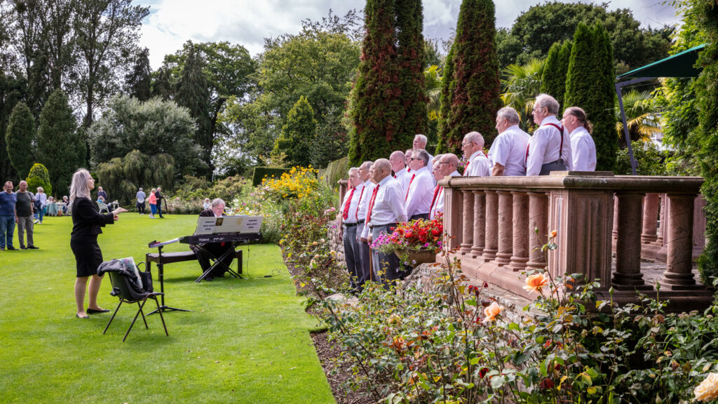 A joyful gathering of individuals in white shirts singing harmoniously amidst a picturesque garden.