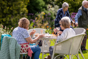 Three elderly people enjoying each other's company at a garden table, surrounded by lush greenery.