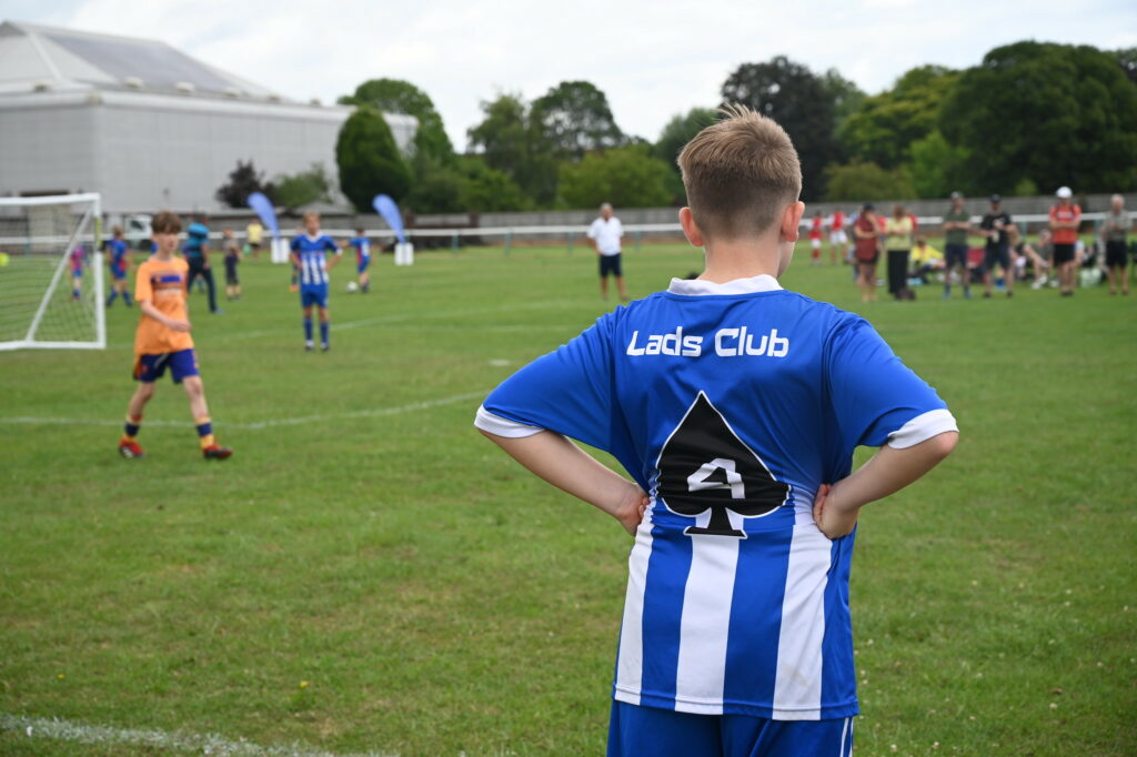 Child playing football with their back to camera stood pitch-side