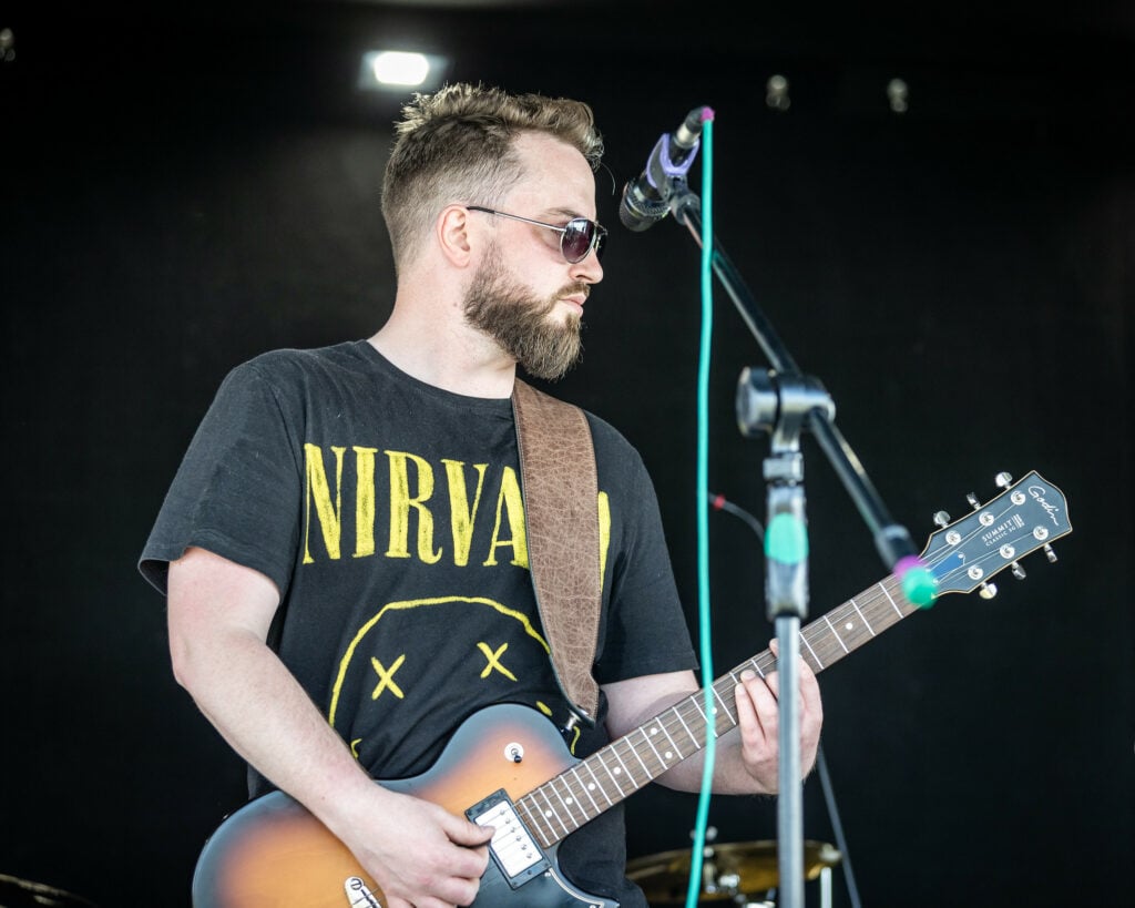 An image of a man with a beard and sunglasses performing on an electric guitar.